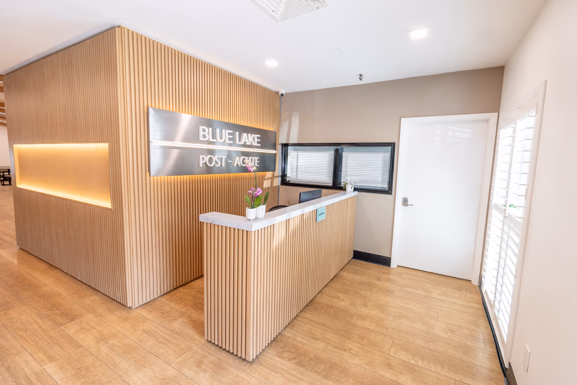 Reception area of Blue Lake Post Acute facility with a modern wooden reception desk and wall paneling, a metallic sign displaying the facility name, a small potted plant on the desk, and a white door and window with blinds in the background.