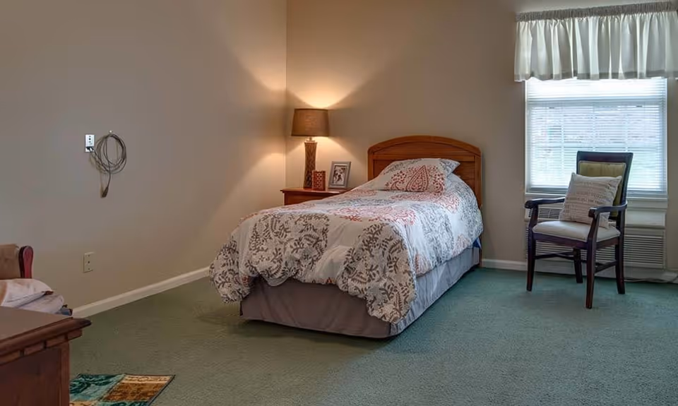 A simple bedroom with a single bed covered in a patterned quilt, a wooden nightstand with a lamp and framed photo, a wooden chair with a cushion near a window with blinds and a valance, and green carpeted floor.