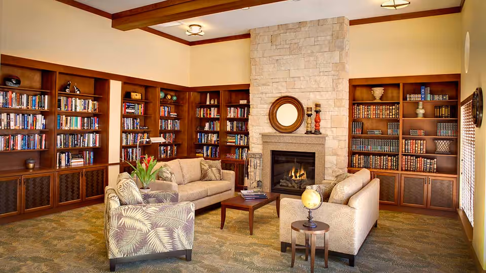 A cozy living room with beige sofas and an armchair arranged around a wooden coffee table. The room features built-in wooden bookshelves filled with books, a stone fireplace with a round mirror above it, and a small side table with a globe. The carpet has a green leafy pattern, and the walls are painted a warm beige color.