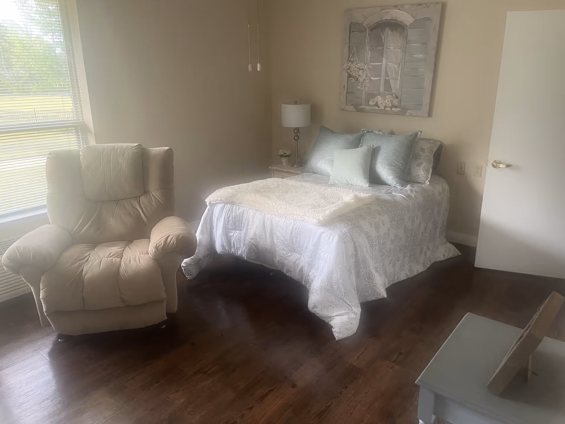 A cozy bedroom with a bed covered in white and light blue bedding, a beige recliner chair near a window with blinds, a nightstand with a lamp and small decoration, and a piece of wall art above the bed depicting a window with flowers.