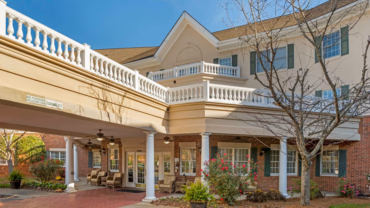Covered entrance/porte-cochère of a brick and stucco senior living building with white columns, outdoor seating, and landscaped shrubs.