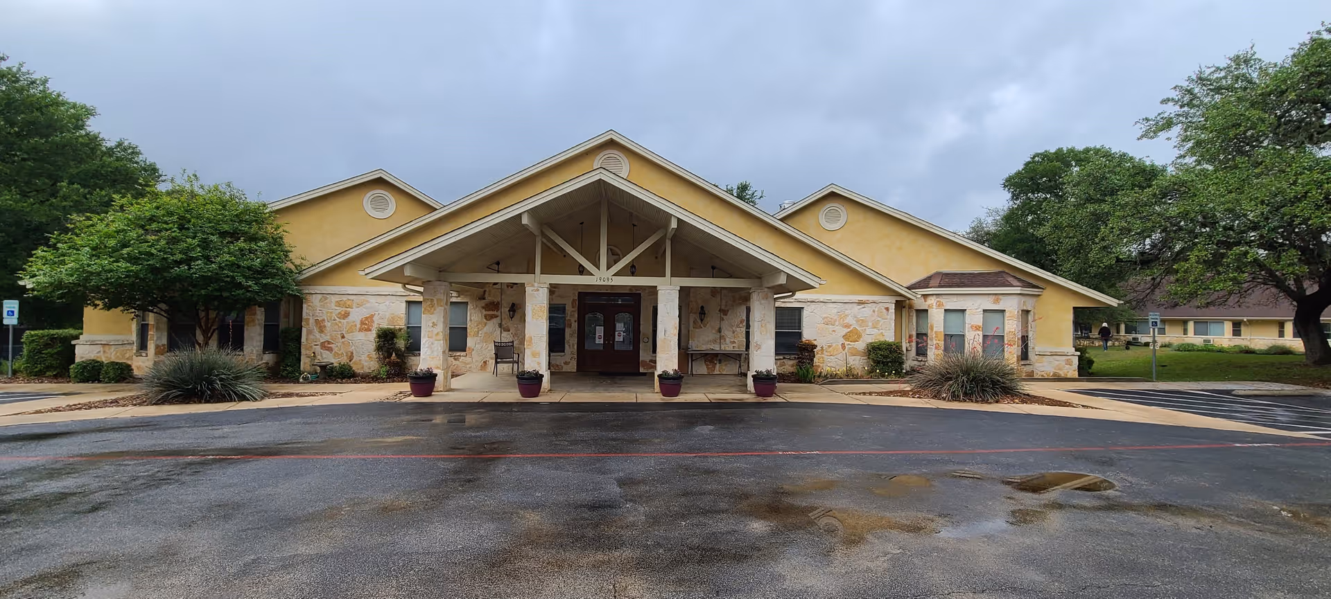 Front entrance of a single-story yellow senior living facility with a covered porte-cochere, stone accents, potted plants, and surrounding trees.