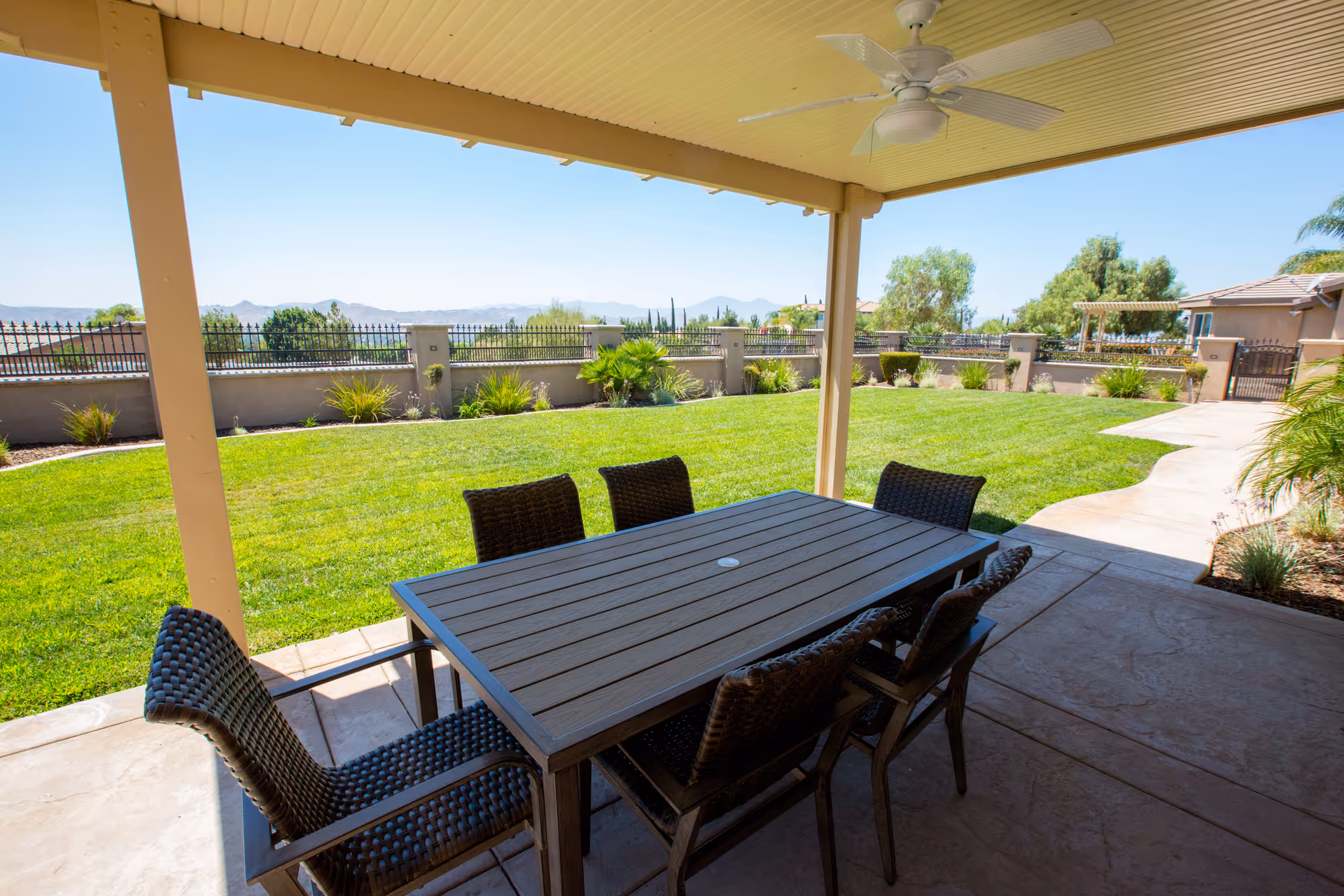 Covered outdoor patio area with a ceiling fan, featuring a rectangular table and six wicker chairs. The patio overlooks a well-maintained grassy yard with various plants along a low wall and a view of distant mountains under a clear blue sky.
