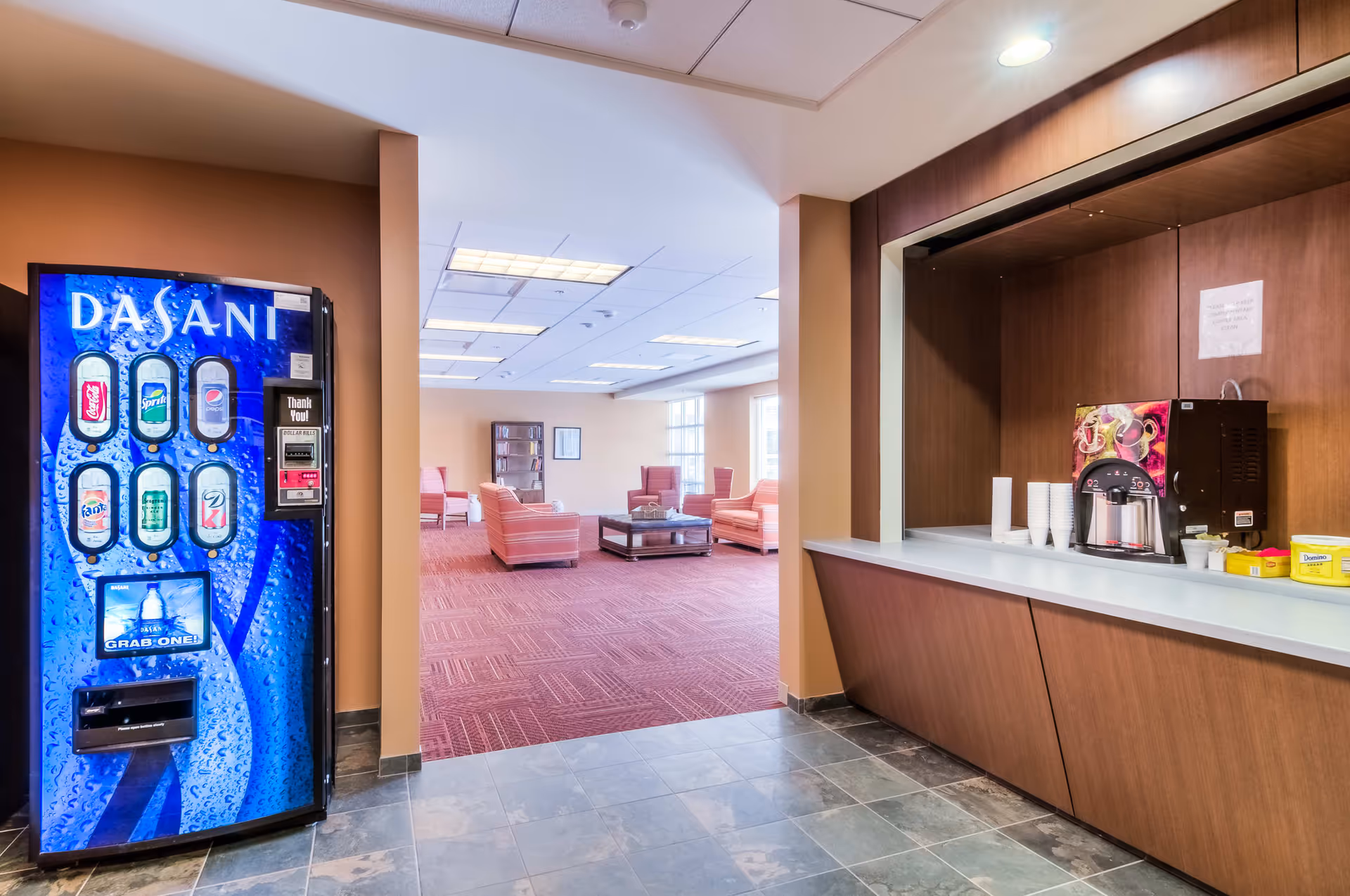 Interior view of a senior living facility common area with a blue Dasani vending machine on the left, a coffee station with cups and condiments on the right, and a lounge area with pink upholstered chairs and a coffee table in the background.