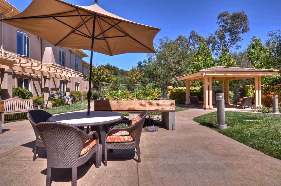 Outdoor patio area at a senior living facility with a round table and four wicker chairs with cushions under a large beige umbrella. There is a wooden bench to the left, a raised garden bed with plants, and a covered seating area with chairs in the background. The area is surrounded by greenery and trees under a clear blue sky.