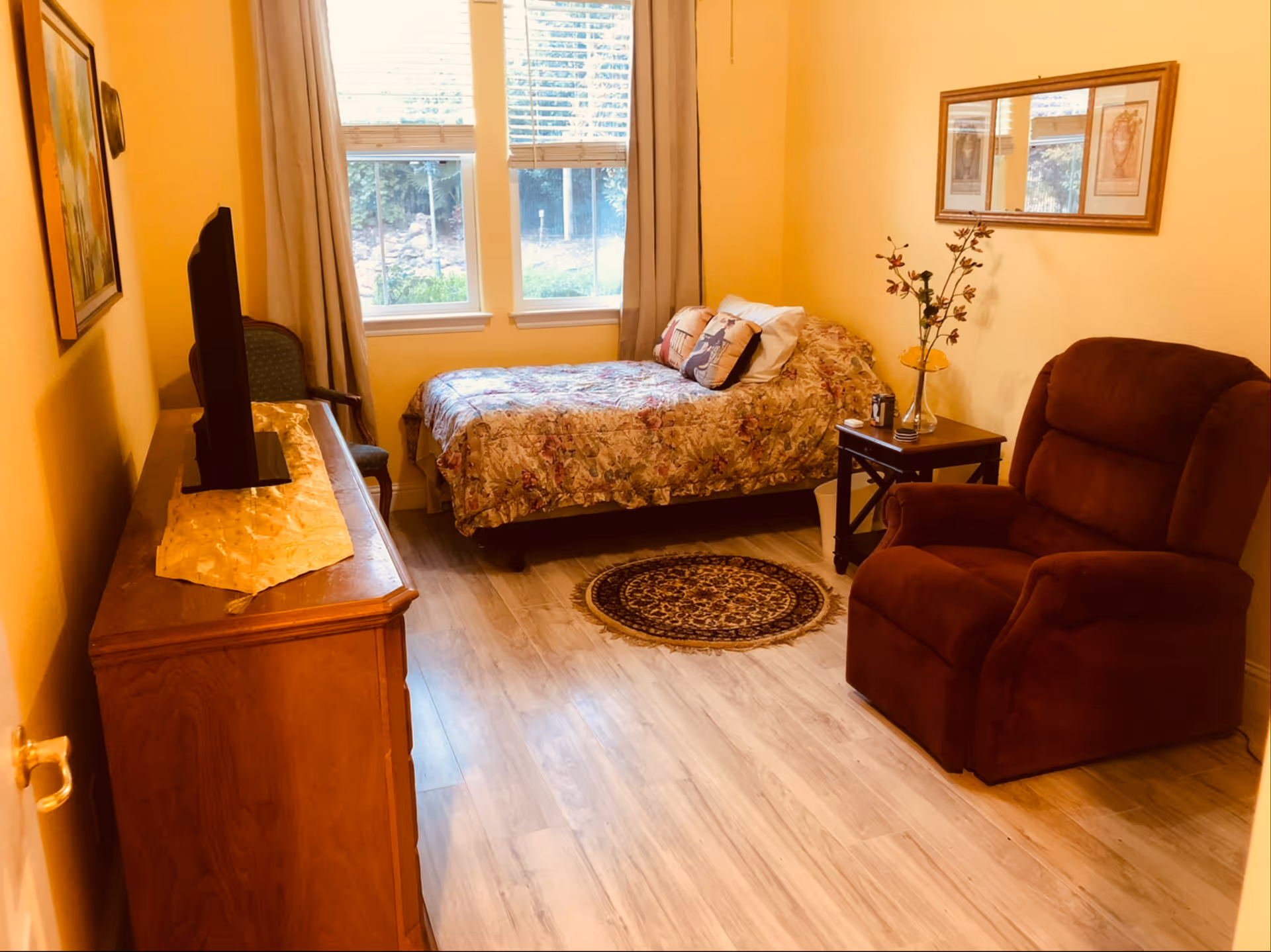 A cozy bedroom with a single bed covered in a floral bedspread, a brown recliner chair, a wooden dresser with a TV on top, a small side table with a vase and decorative branches, a round patterned rug on the floor, and two windows with beige curtains letting in natural light.