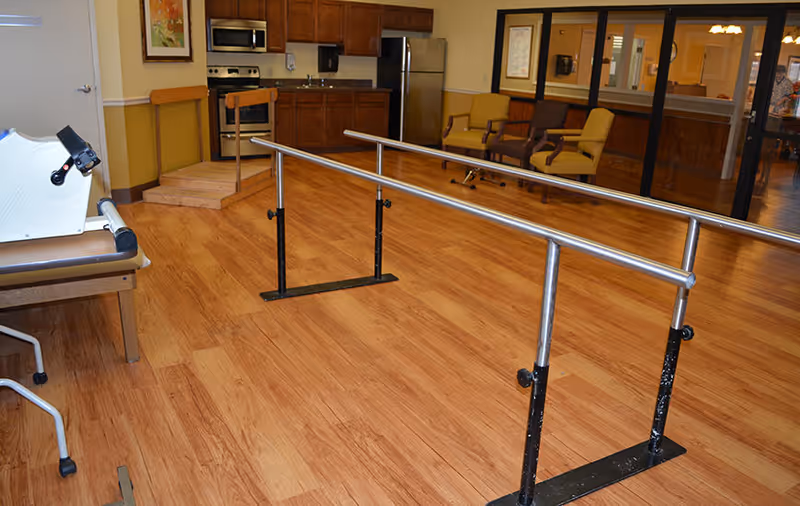 Interior room with parallel walking bars on a wood floor, exercise equipment, a kitchenette and seating in the background.