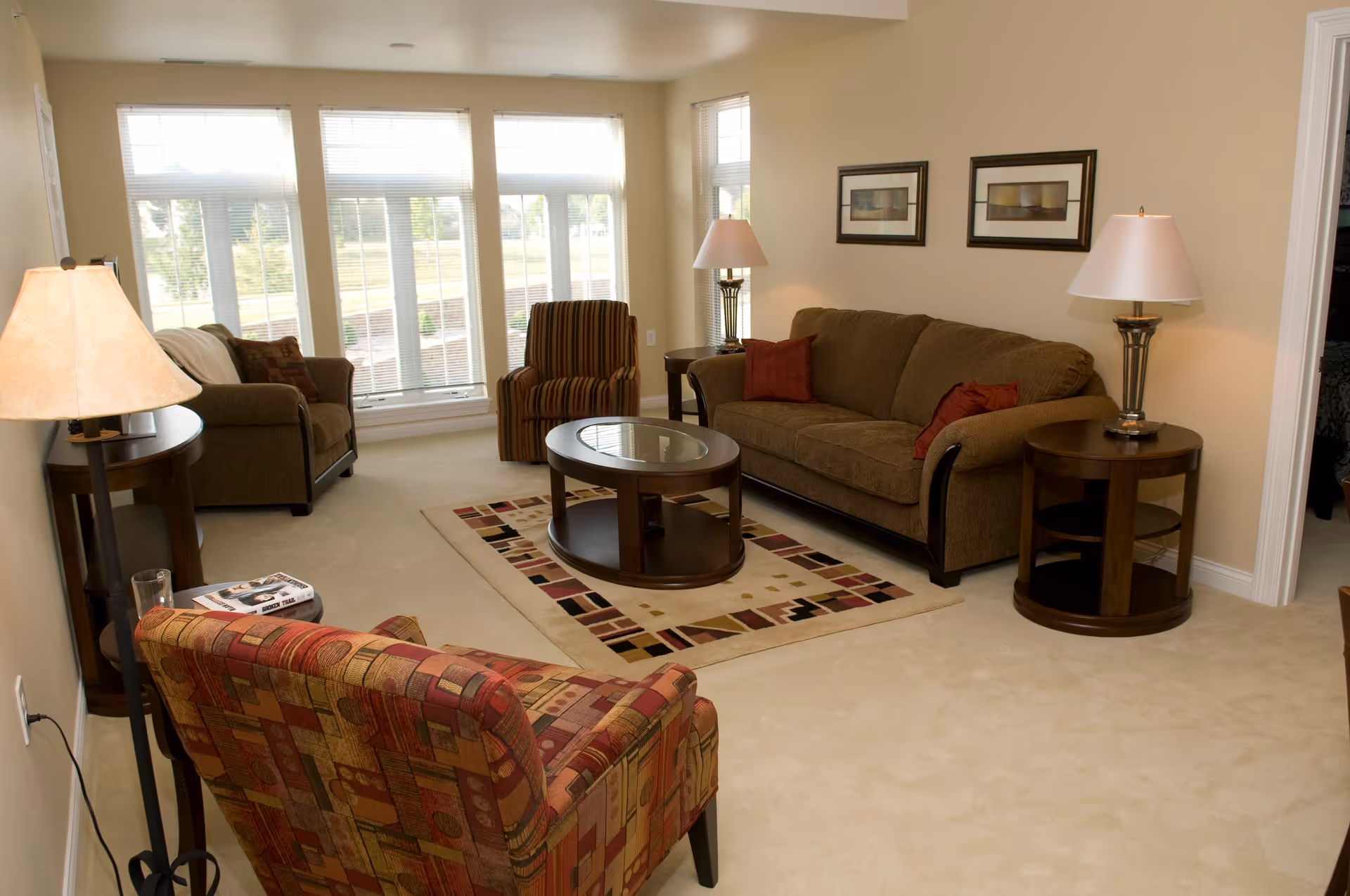 A well-lit living room with beige walls and carpet, featuring a brown sofa with red cushions, a striped armchair, a patterned armchair, a wooden oval coffee table on a decorative rug, two side tables with lamps, and large windows with blinds letting in natural light.