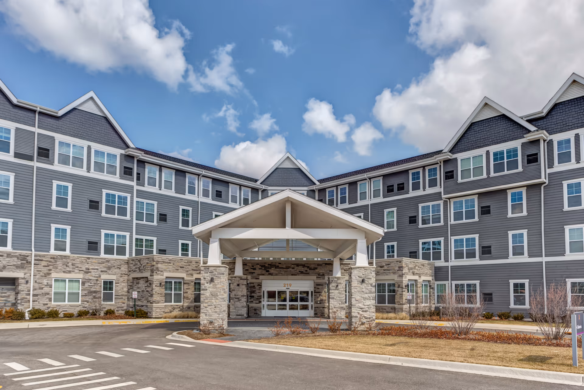 Front exterior view of a large senior living facility building with gray siding and stone accents. The building has multiple windows and a covered entrance with pillars. The sky is partly cloudy with blue patches visible.