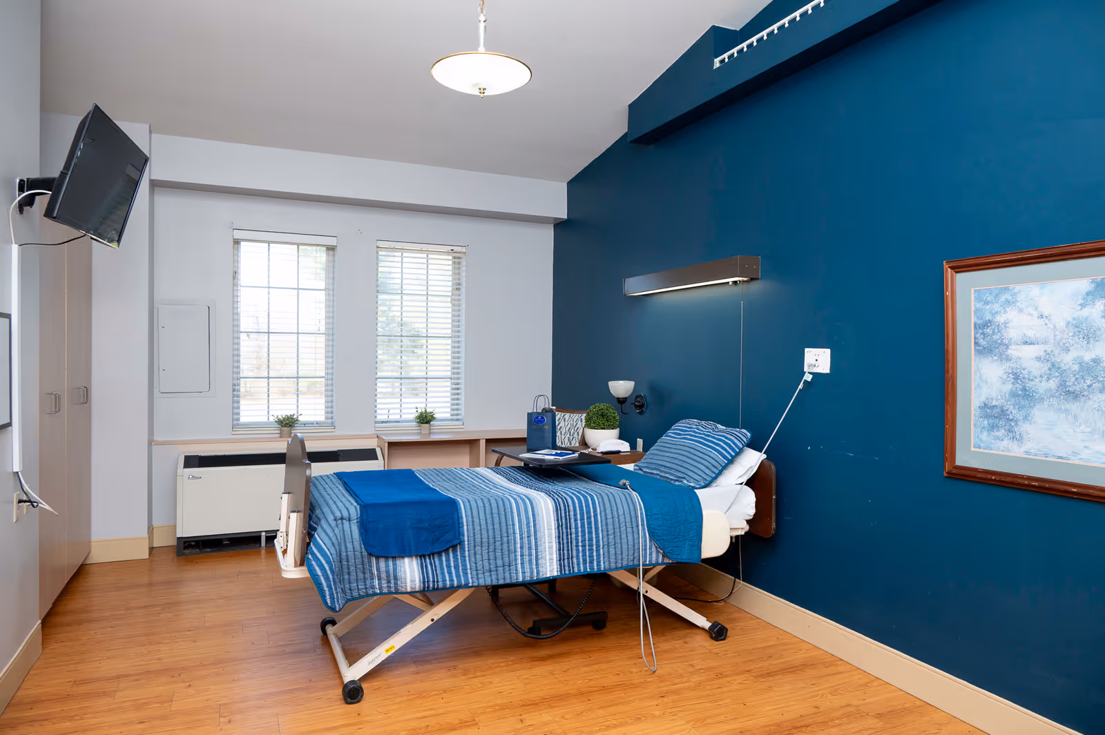 A rehabilitation room with a hospital bed covered in blue striped bedding. The room has wooden flooring, a deep blue accent wall with a framed picture, two windows with white blinds, a wall-mounted TV, and a small table with a lamp and decorative plants.