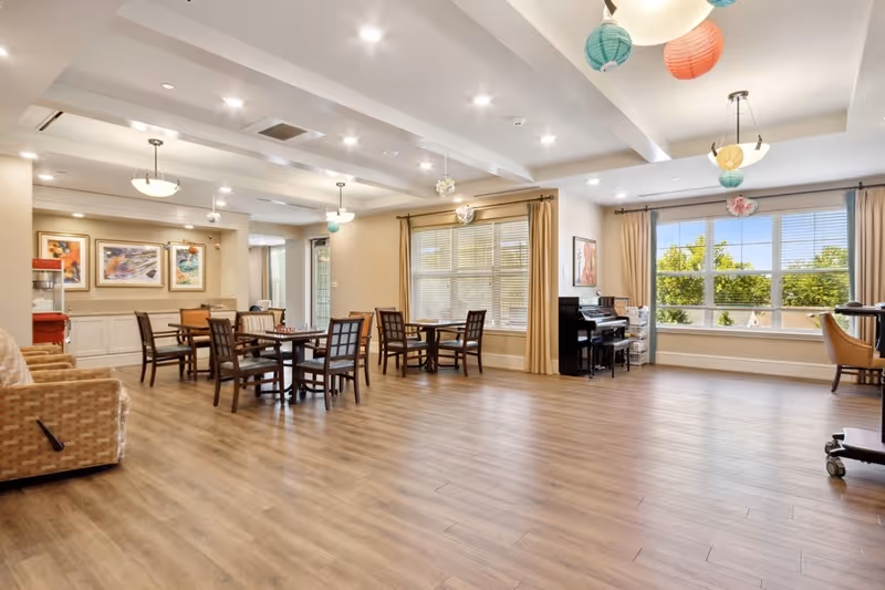 A spacious, well-lit common area with wooden flooring, several tables and chairs arranged for seating, large windows with beige curtains letting in natural light, a piano against the wall, and colorful hanging paper lantern decorations from the ceiling.