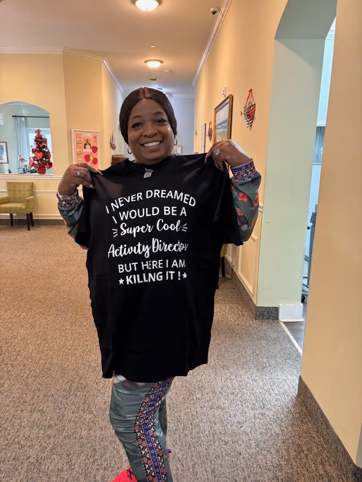 A smiling woman standing in a hallway of an assisted living facility holding up a black t-shirt that reads 'I never dreamed I would be a super cool activity director but here I am killing it!'. The hallway has beige walls, carpeted floor, and some framed pictures and decorations on the walls.