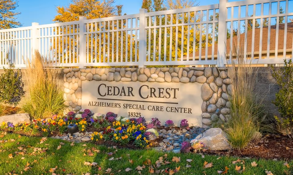 Stone sign for Cedar Crest Alzheimer's Special Care Center surrounded by colorful flowers and greenery, with a white fence and trees in the background under a clear blue sky.