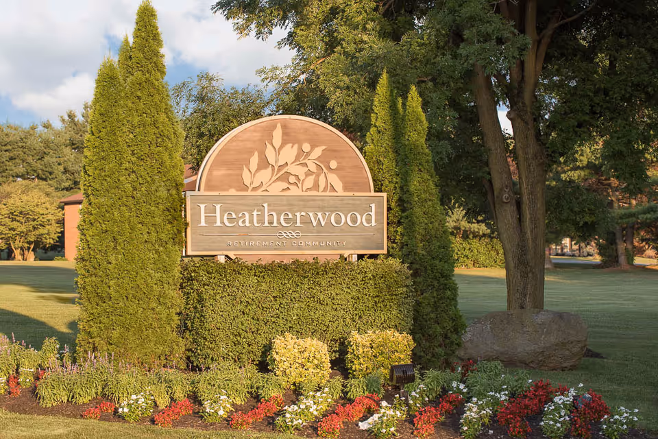 A wooden sign for Heatherwood Retirement Community surrounded by neatly trimmed bushes, colorful flowers, and tall evergreen trees, set in a well-maintained grassy area with larger trees in the background under a partly cloudy sky.