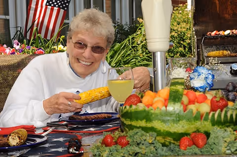 An elderly woman wearing sunglasses and a white sweater is smiling while holding a piece of grilled corn on the cob at an outdoor table. The table is decorated with a patriotic tablecloth and has a large carved watermelon bowl filled with various fruits, including strawberries, cantaloupe, and grapes. An American flag is visible in the background along with greenery and a grill with food cooking on it.