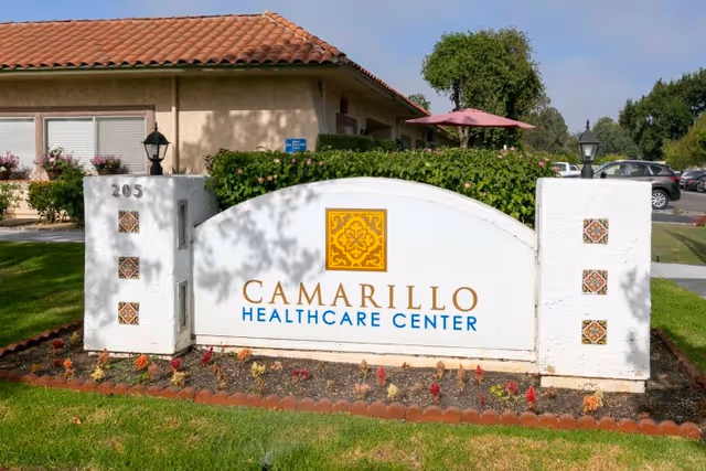 Outdoor view of the entrance sign for Camarillo Healthcare Center, featuring a white stucco sign with decorative tiles and landscaping, with part of the building and parking lot visible in the background.