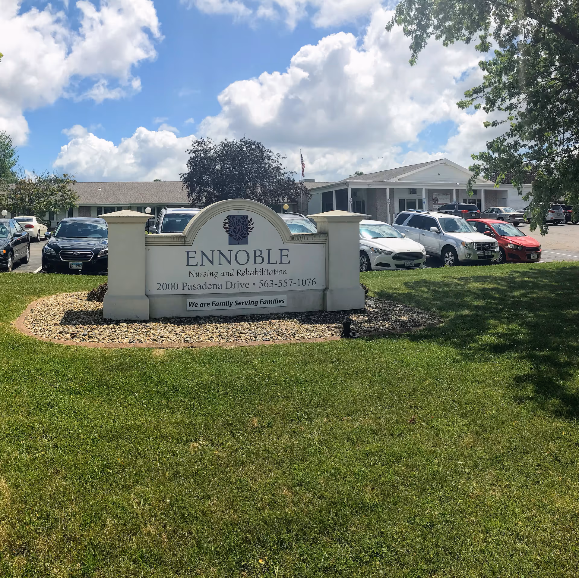 Outdoor view of Ennoble Nursing and Rehabilitation facility sign with the building and parked cars in the background under a partly cloudy sky.