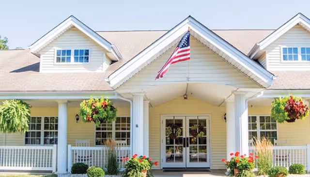 Front exterior view of a building with a beige facade, white columns, and a peaked roof. An American flag is mounted above the entrance. There are hanging plants and potted flowers decorating the porch area.
