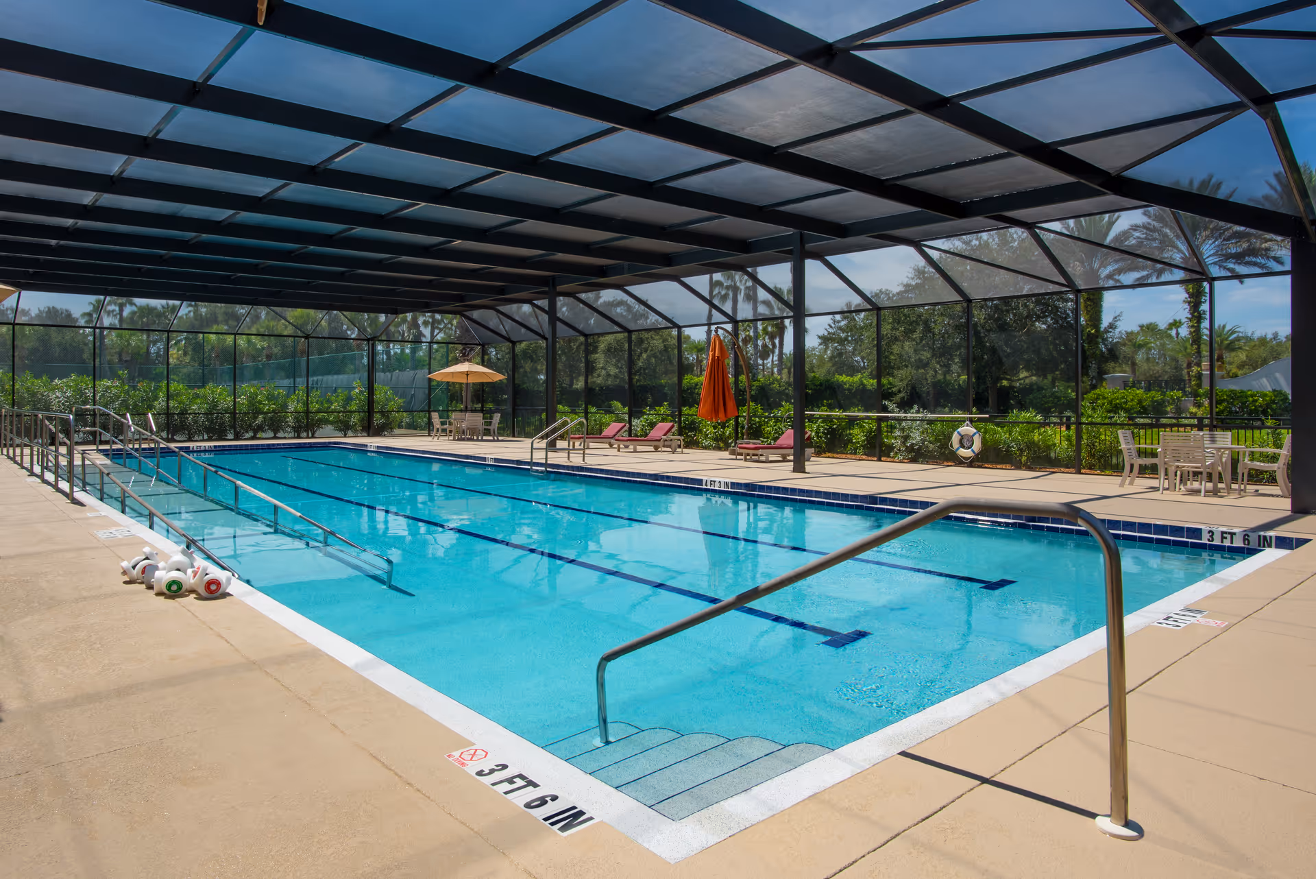 An enclosed outdoor swimming pool with lane markings, handrails, lounge chairs and umbrellas under a screened roof.