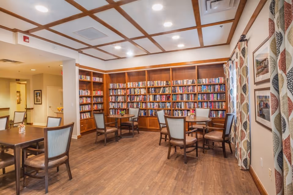 A cozy reading room with wooden floors and ceiling beams, featuring multiple round tables with chairs arranged around them. The back wall is lined with built-in wooden bookshelves filled with books. The room is well-lit with recessed ceiling lights and has patterned curtains on the right side covering windows. Framed pictures hang on the wall next to the windows.