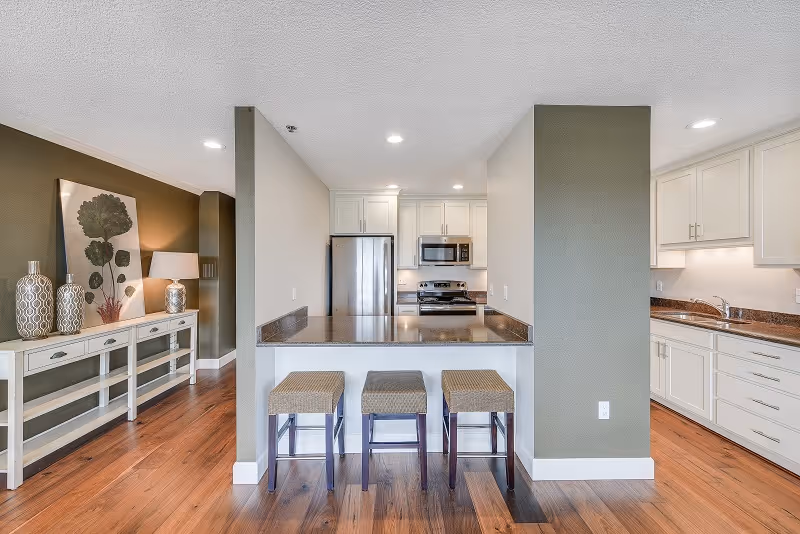 Open-concept kitchen with a breakfast bar and three stools, stainless steel appliances, white cabinets, and hardwood floors.