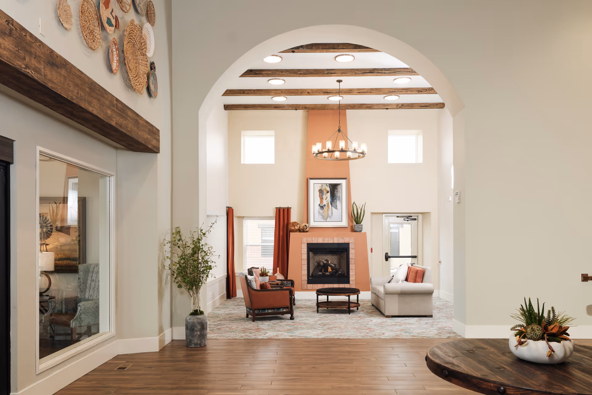 Spacious senior living lounge with an arched entry, fireplace and artwork, seating area, wooden ceiling beams and decorative wall baskets.