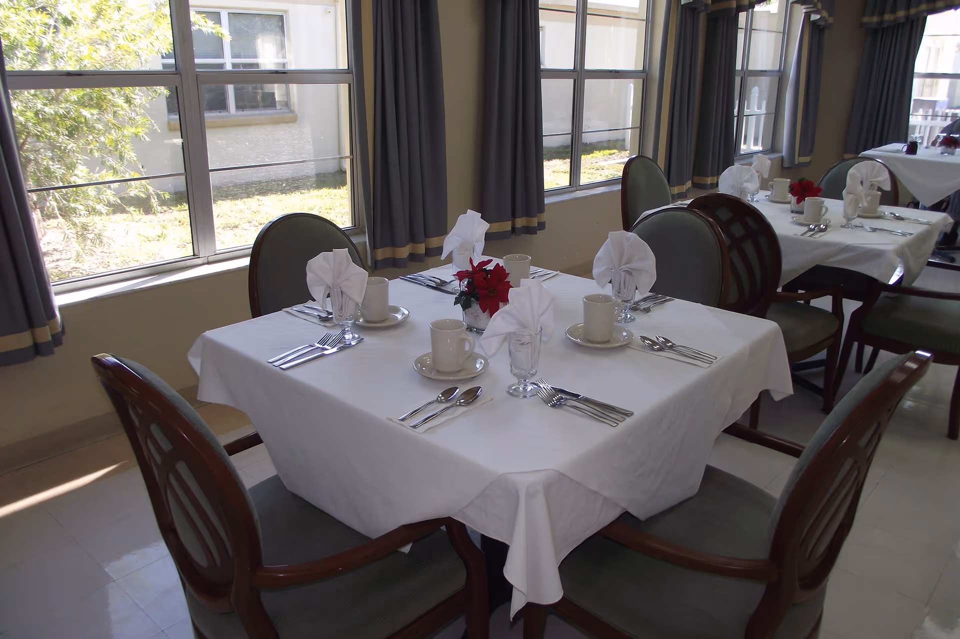 A dining room with tables set for four people, each table covered with a white tablecloth, napkins folded in a decorative manner, cups, glasses, and silverware. The room has large windows with gray curtains allowing natural light to enter.