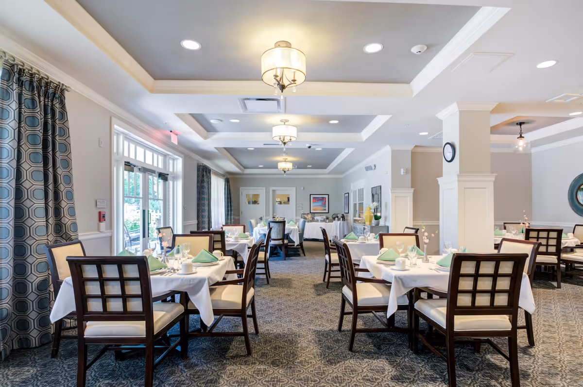 A bright and elegant dining room with multiple tables set with white tablecloths, green napkins, glassware, and cups. The room features patterned carpet, large windows with geometric patterned curtains, and ceiling lights with decorative fixtures. There is a buffet table at the far end of the room.