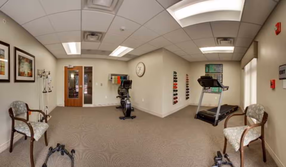 A small fitness room in a senior living facility with exercise equipment including a treadmill, a stationary bike, and a rack of colorful dumbbells. The room has beige walls, carpeted floor, two chairs with patterned upholstery, framed artwork on the walls, and a clock. There is a wooden door with glass panels and an exit sign on the wall.