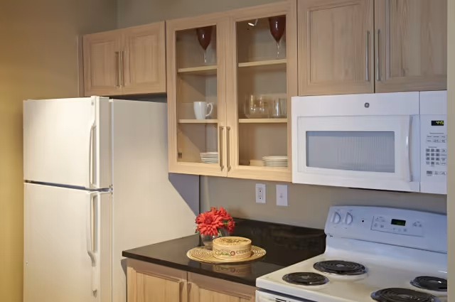 A kitchen area with light wood cabinets, a white refrigerator, a white microwave mounted above a white electric stove, and a black countertop with a small decorative tray and red flowers.