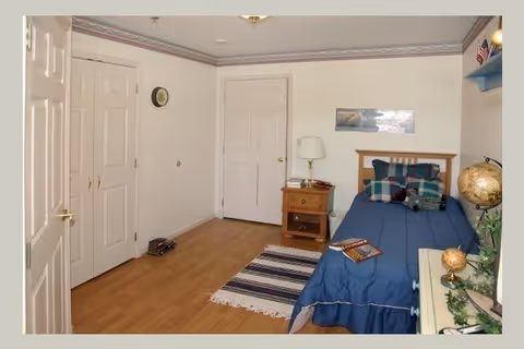 A small bedroom with a single bed covered in a blue bedspread and several plaid pillows. Next to the bed is a wooden nightstand with a lamp and some books. The room has light-colored walls, a wooden floor, a striped rug, and a globe on a dresser. There are two closed white doors and a clock on the wall.