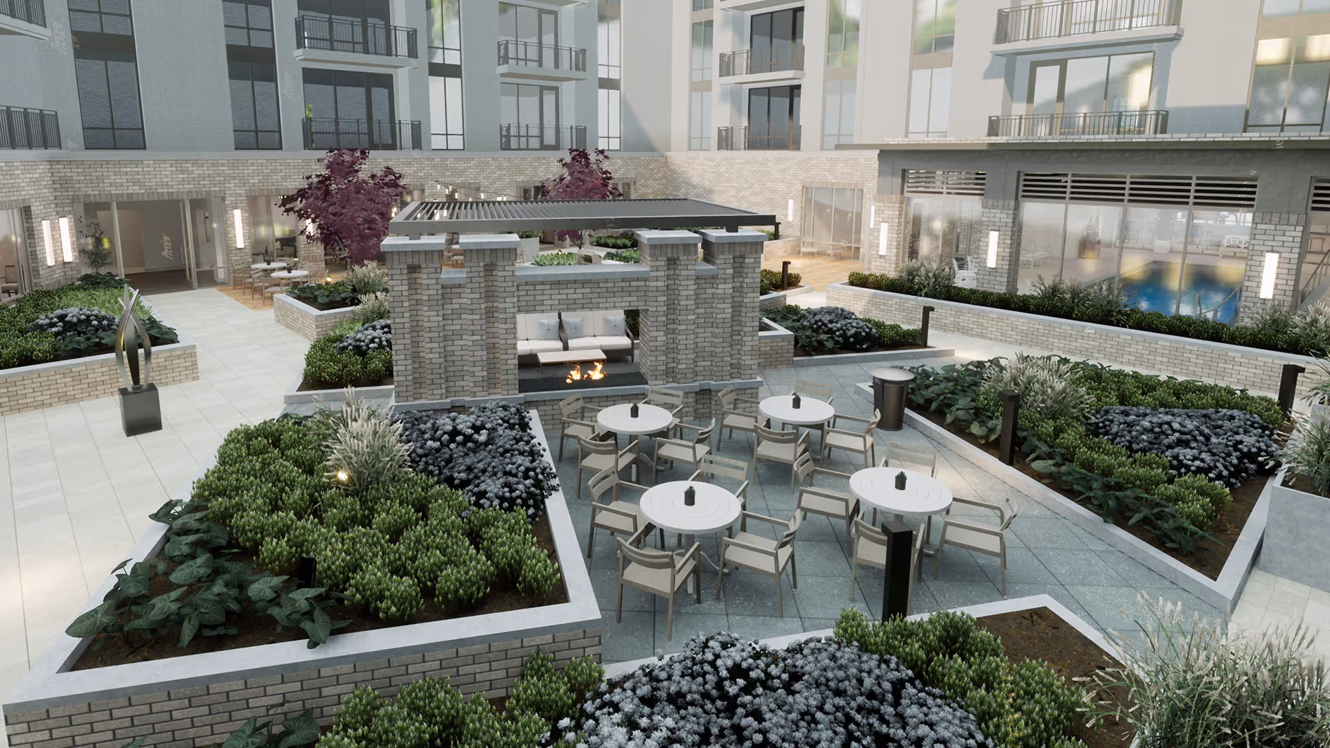 Outdoor courtyard area with multiple round tables and chairs arranged around a central brick fireplace. The space is surrounded by raised garden beds filled with various green plants and shrubs. The courtyard is part of a multi-story building with large windows and balconies overlooking the area.