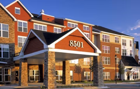 Exterior view of a senior living facility building with a covered entrance displaying the number 8501. The building has multiple windows and a combination of brick and stone facade under a clear blue sky.