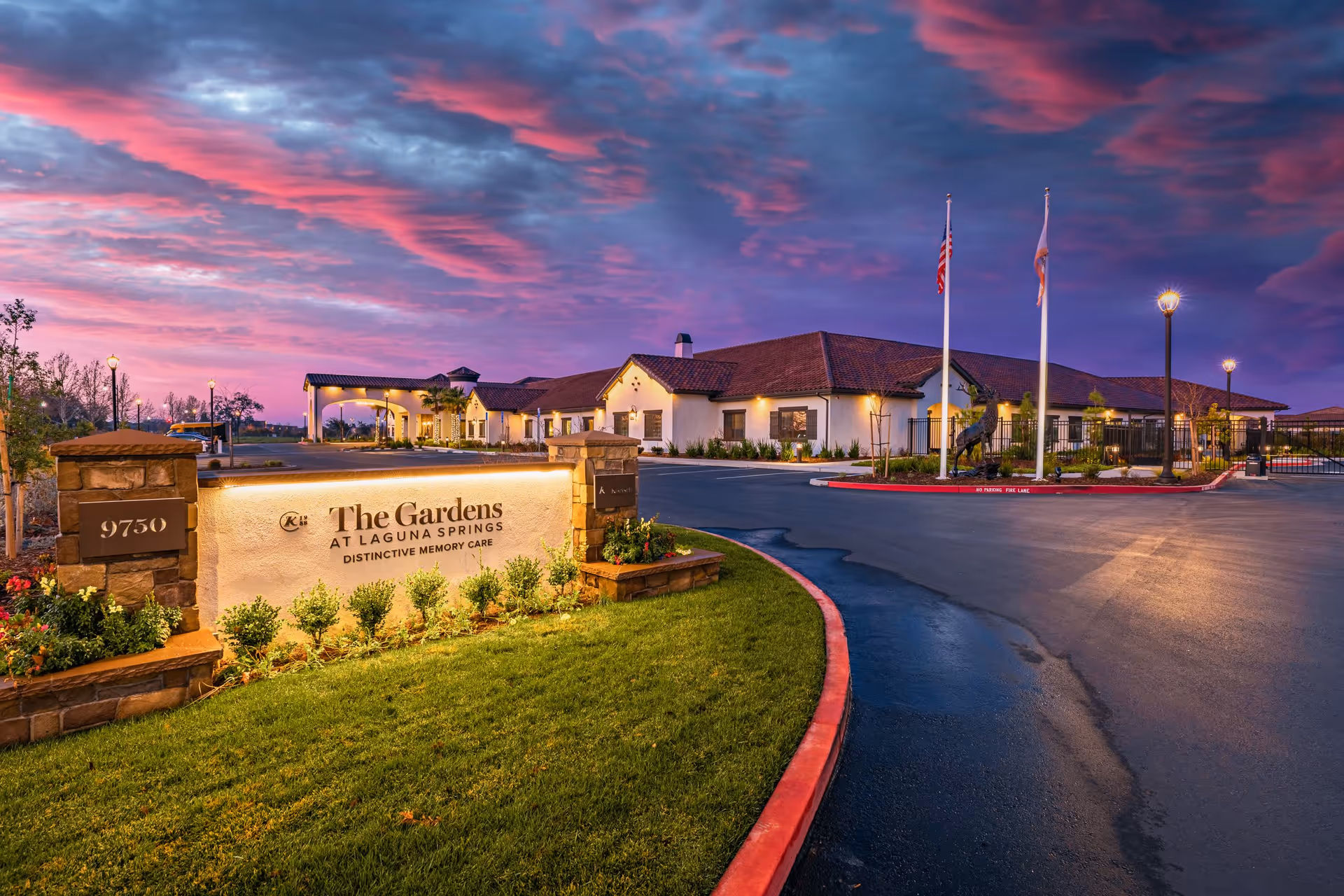 Exterior view of The Gardens At Laguna Springs facility at dusk with a dramatic purple and pink sky. The building is single-story with a tiled roof, surrounded by a paved driveway and landscaped greenery. Two flagpoles with flags and a statue are visible near the entrance.