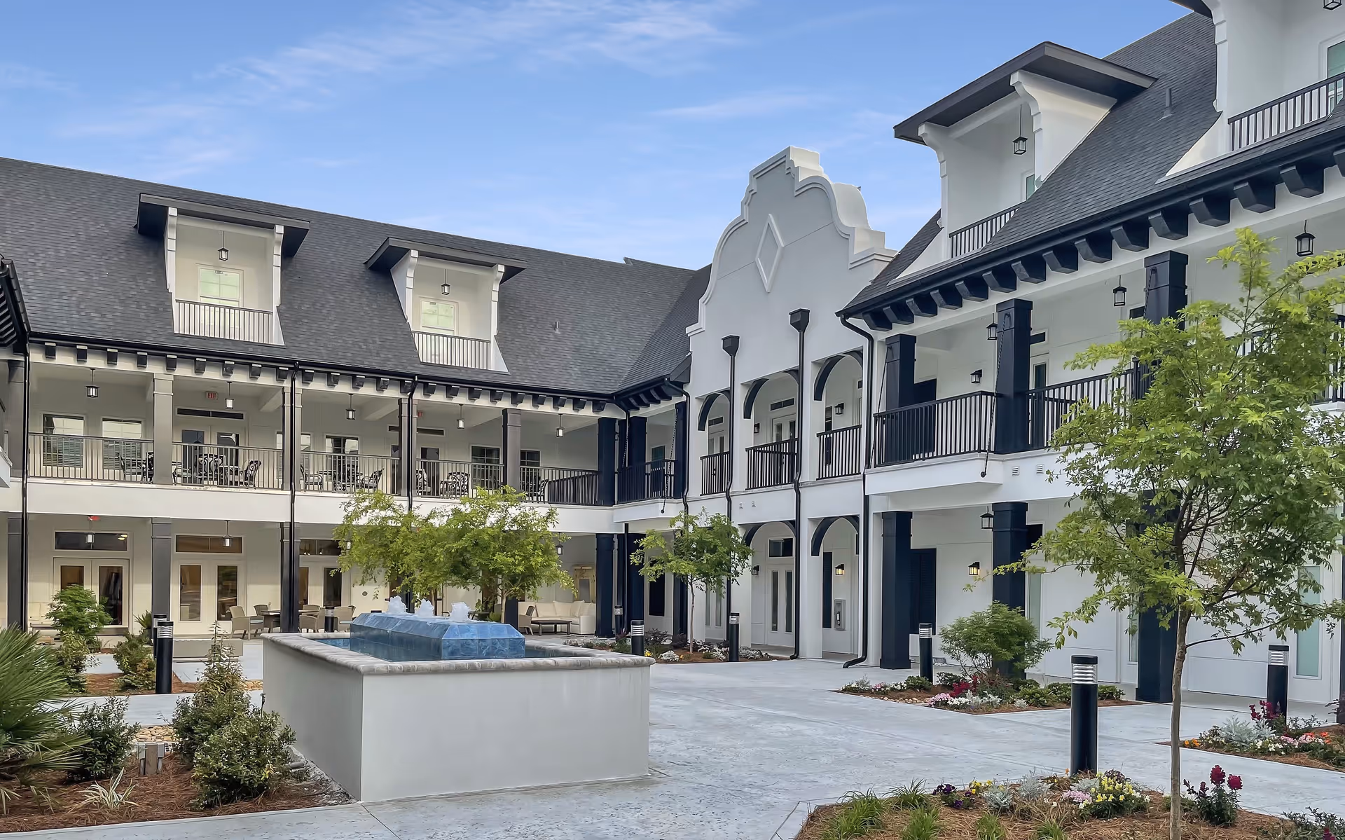 Outdoor courtyard area of The Blake at Miramar Beach senior living facility featuring a central water fountain, landscaped garden beds with small trees and flowers, and a two-story building with balconies and covered walkways surrounding the courtyard under a clear blue sky.