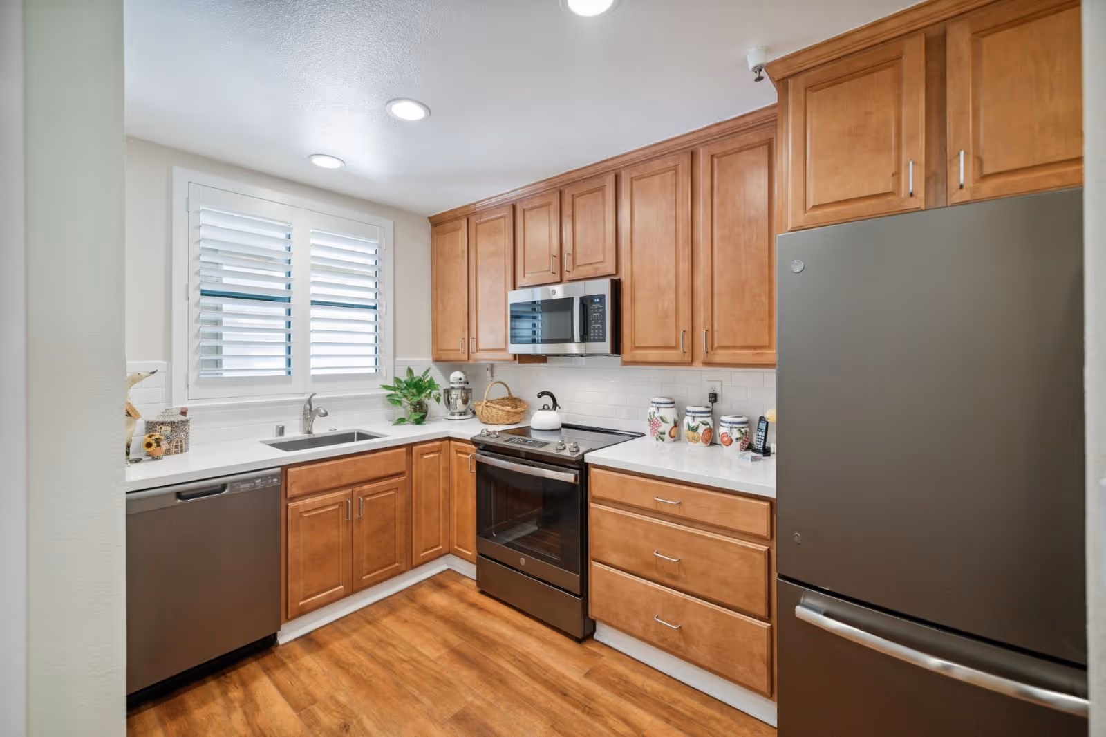 A modern kitchen with wooden cabinets, white countertops, and stainless steel appliances including a refrigerator, dishwasher, oven, and microwave. There is a window with white shutters above the sink, and decorative jars and a plant on the countertop.