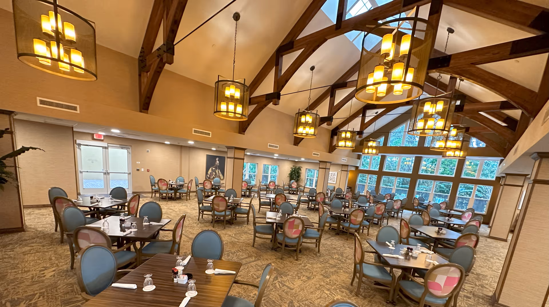 Spacious dining room with multiple tables and chairs arranged neatly. The room features high vaulted ceilings with exposed wooden beams and large hanging light fixtures. Large windows on one side allow natural light to fill the space, and the walls are decorated with artwork and plants.