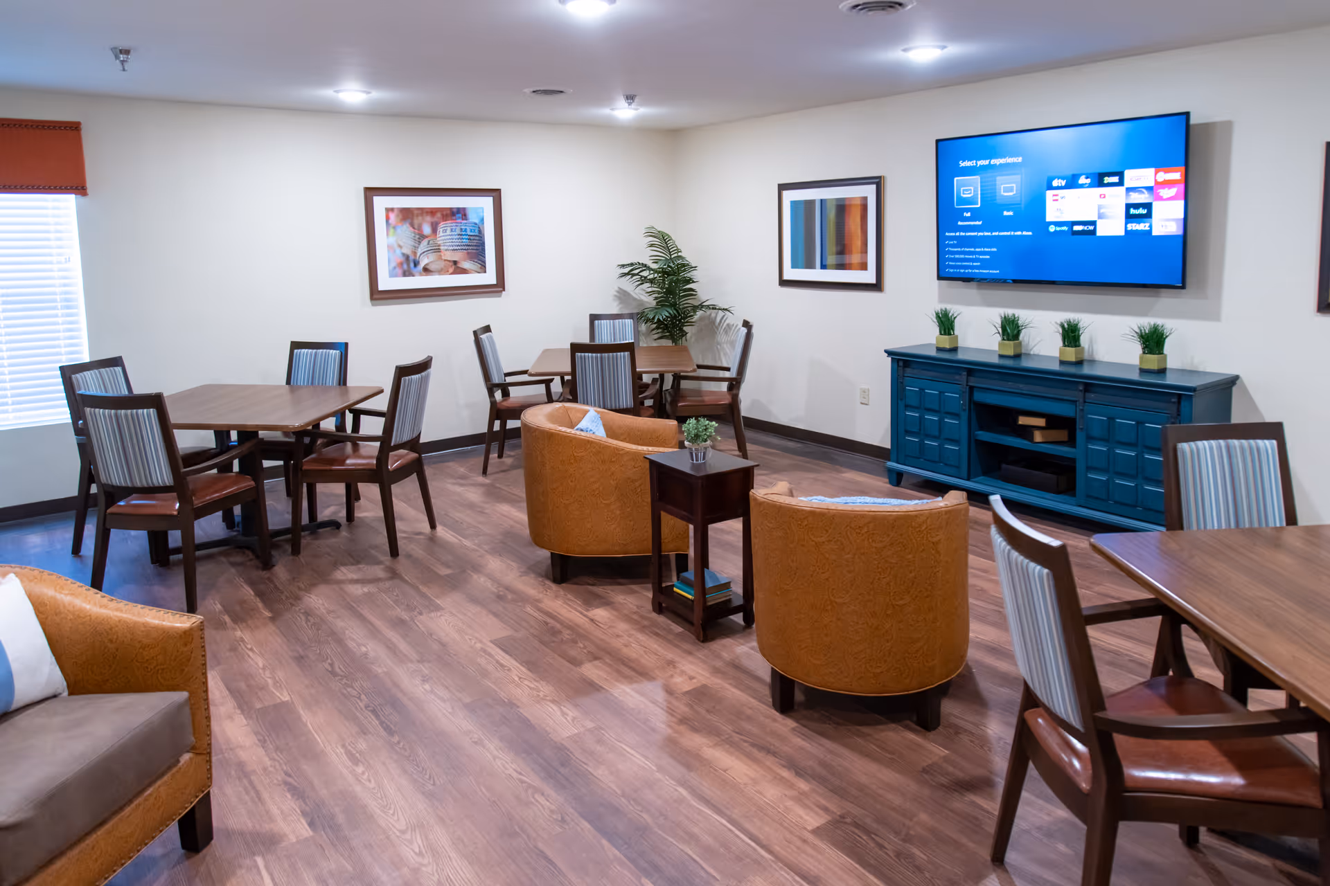A well-lit common area with wooden flooring featuring several tables and chairs arranged for seating. Two comfortable brown armchairs face a blue console table with small potted plants on top and a large flat-screen TV mounted on the wall above it. The walls are decorated with framed artwork, and a window with a red valance allows natural light into the room.