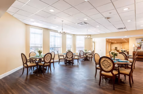 Bright dining room with multiple round tables and upholstered chairs, large windows and chandeliers.