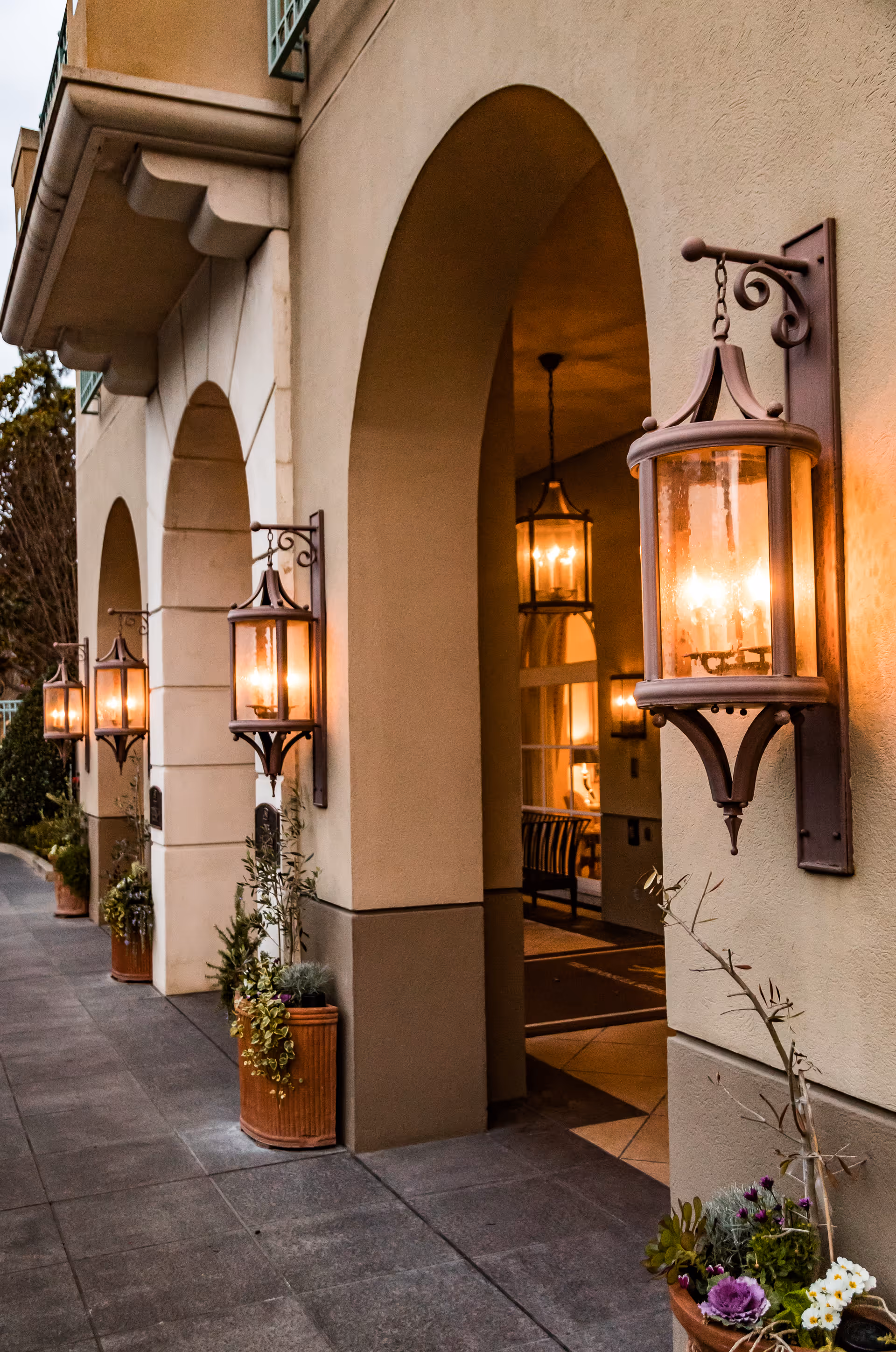 Exterior view of a building with arched doorways and wall-mounted lantern-style lights glowing warmly. Several potted plants are placed along the walkway beside the building.