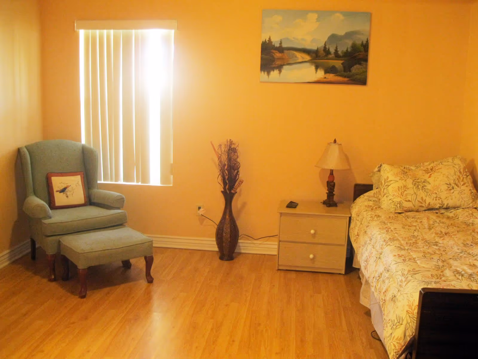 A cozy bedroom with a wooden floor, a floral-patterned bed on the right, a wooden nightstand with a lamp and remote control, a decorative vase with dried branches in the center, and a green upholstered armchair with an ottoman and a bird-themed pillow near a window with vertical blinds. A landscape painting hangs on the wall above the nightstand.