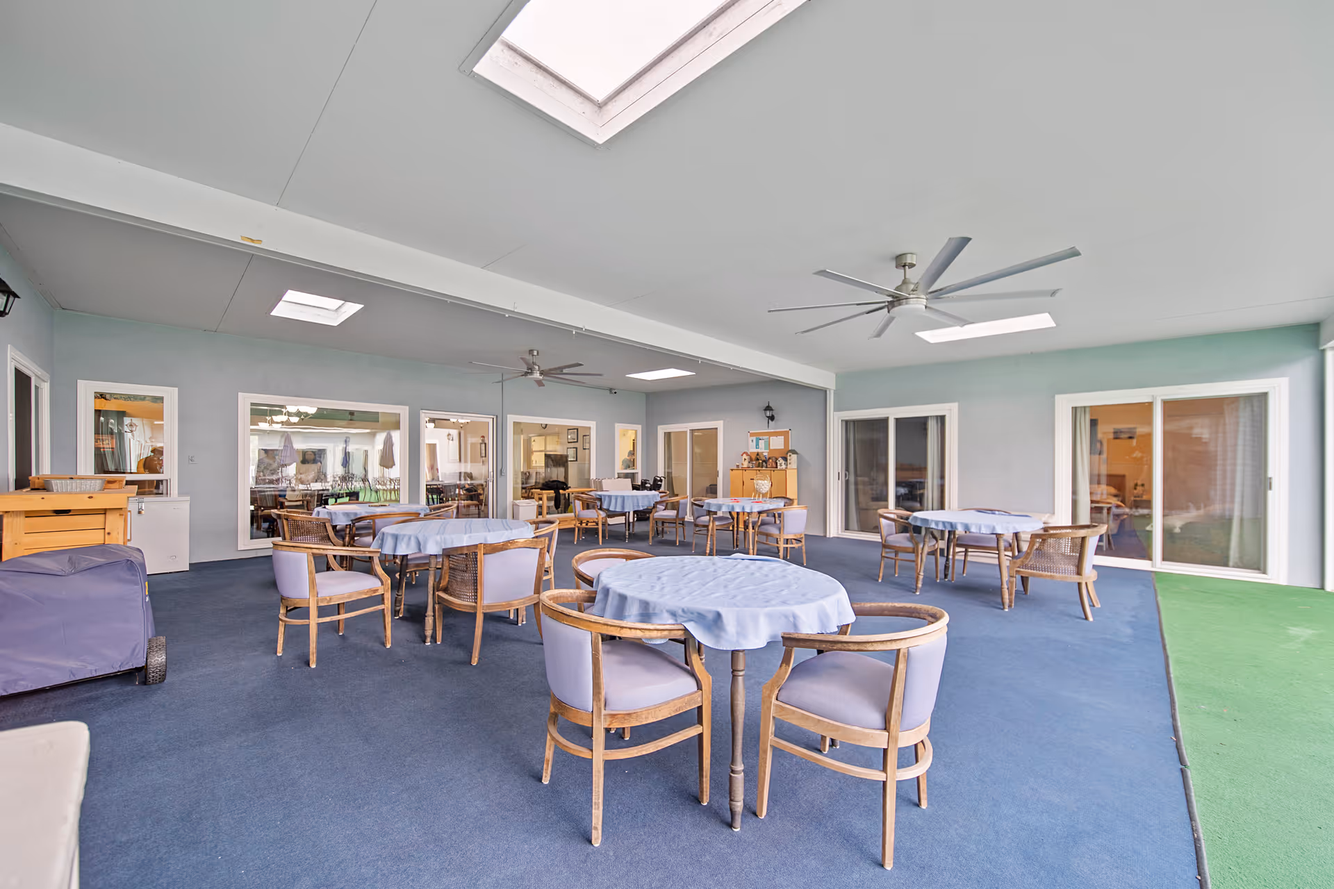 Indoor common area with multiple round tables covered with light blue tablecloths and surrounded by wooden chairs with light purple cushions. The room has a blue carpet, light blue walls, ceiling fans, and skylights. Large windows and sliding glass doors provide views into adjacent rooms.