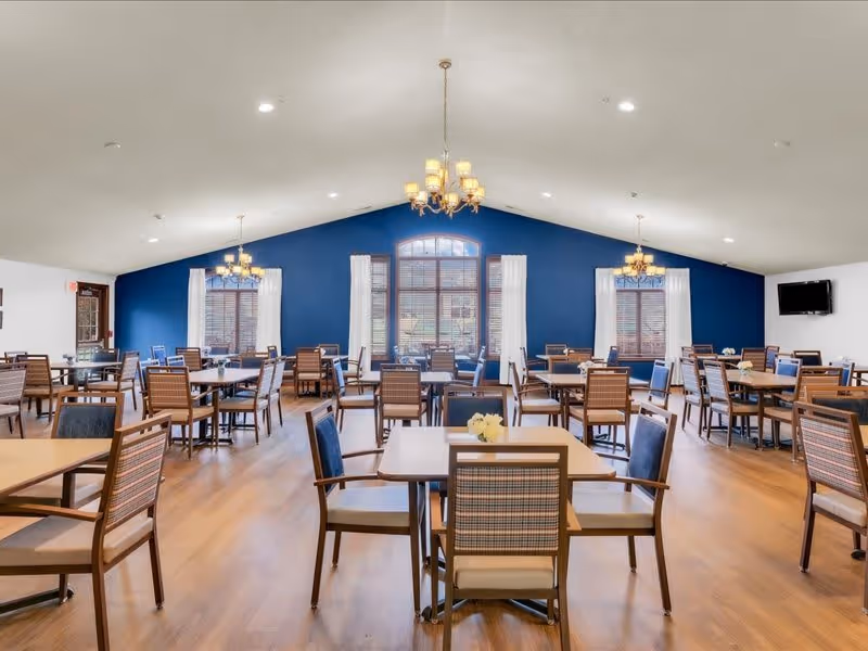A spacious dining room with multiple wooden tables and chairs arranged neatly. The room features a high ceiling with three chandeliers, large windows with white curtains, and a blue accent wall at the far end. The floor is wooden, and there is a wall-mounted television on the right side.
