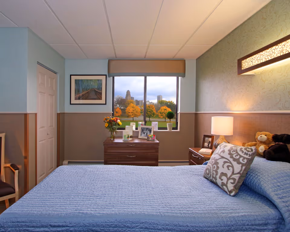 A tidy bedroom with a blue quilted bed, decorative pillows, a nightstand and dresser beneath a window overlooking trees and city buildings.
