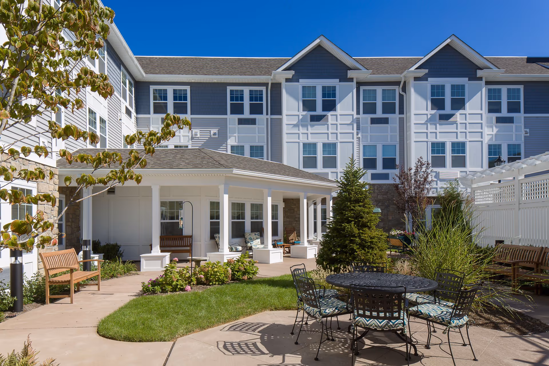 Outdoor courtyard area of a senior living facility with a round metal table and six cushioned chairs on a paved patio. Surrounding the patio are green grass, plants, and trees. The building in the background is three stories tall with white and gray siding, multiple windows, and a covered porch with wooden benches and cushioned chairs.