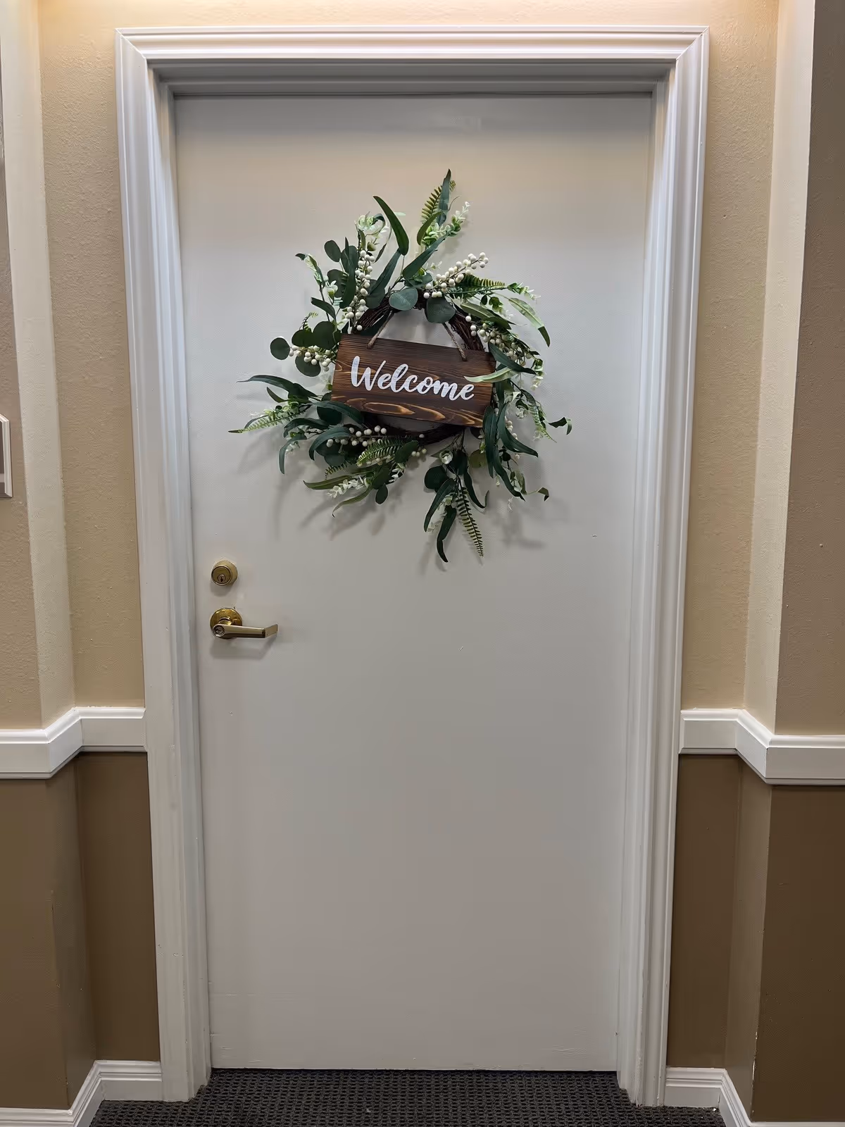 A closed white door with a brass handle and lock, decorated with a green leafy wreath that has white berries and a wooden sign in the center reading 'Welcome'. The door is framed with white trim and the surrounding walls are painted beige with a white chair rail and brown lower section. The floor is carpeted in a dark pattern.
