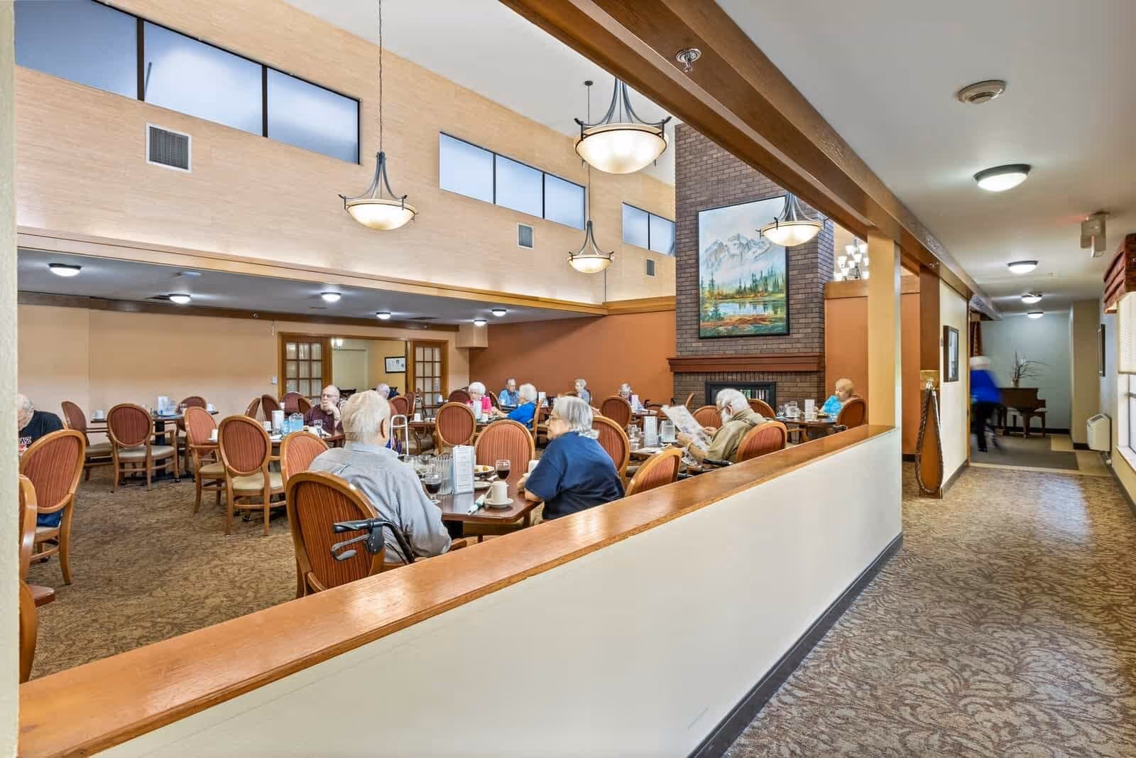 A dining room in a retirement home with elderly residents seated at tables, eating and socializing. The room has high ceilings with hanging light fixtures, a large brick fireplace with a landscape painting above it, and a carpeted floor. There is a hallway to the right with a person walking and a piano at the end.