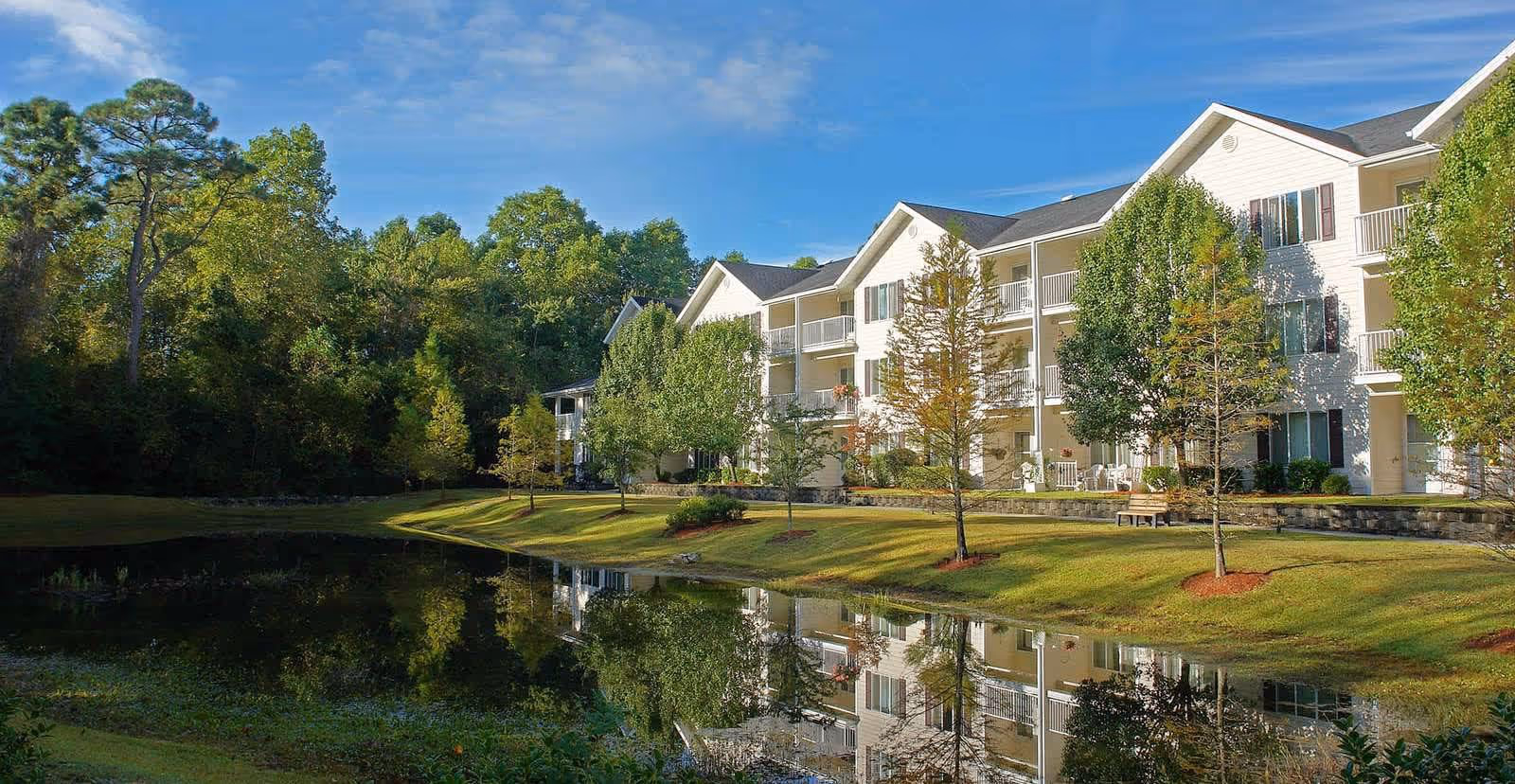 A multi-story residential building with balconies overlooking a calm pond, surrounded by green trees and a well-maintained lawn under a blue sky.