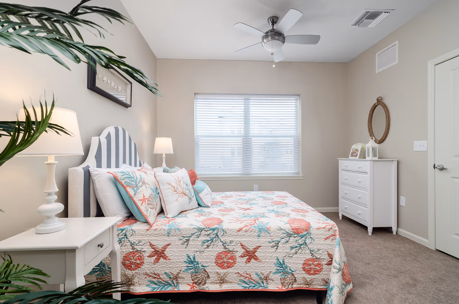 A bright bedroom with a bed covered in a colorful seashell and starfish patterned quilt and matching pillows. The bed has a striped headboard and is flanked by two white bedside tables with lamps. A white dresser with a round mirror above it is against the wall, and a large window with blinds lets in natural light. A ceiling fan is mounted on the ceiling, and a green plant is partially visible in the foreground.