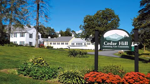 Well-kept lawn and flowerbeds lead to a white multi-wing building with a large 'Cedar Hill Continuing Care Community' sign in the foreground.